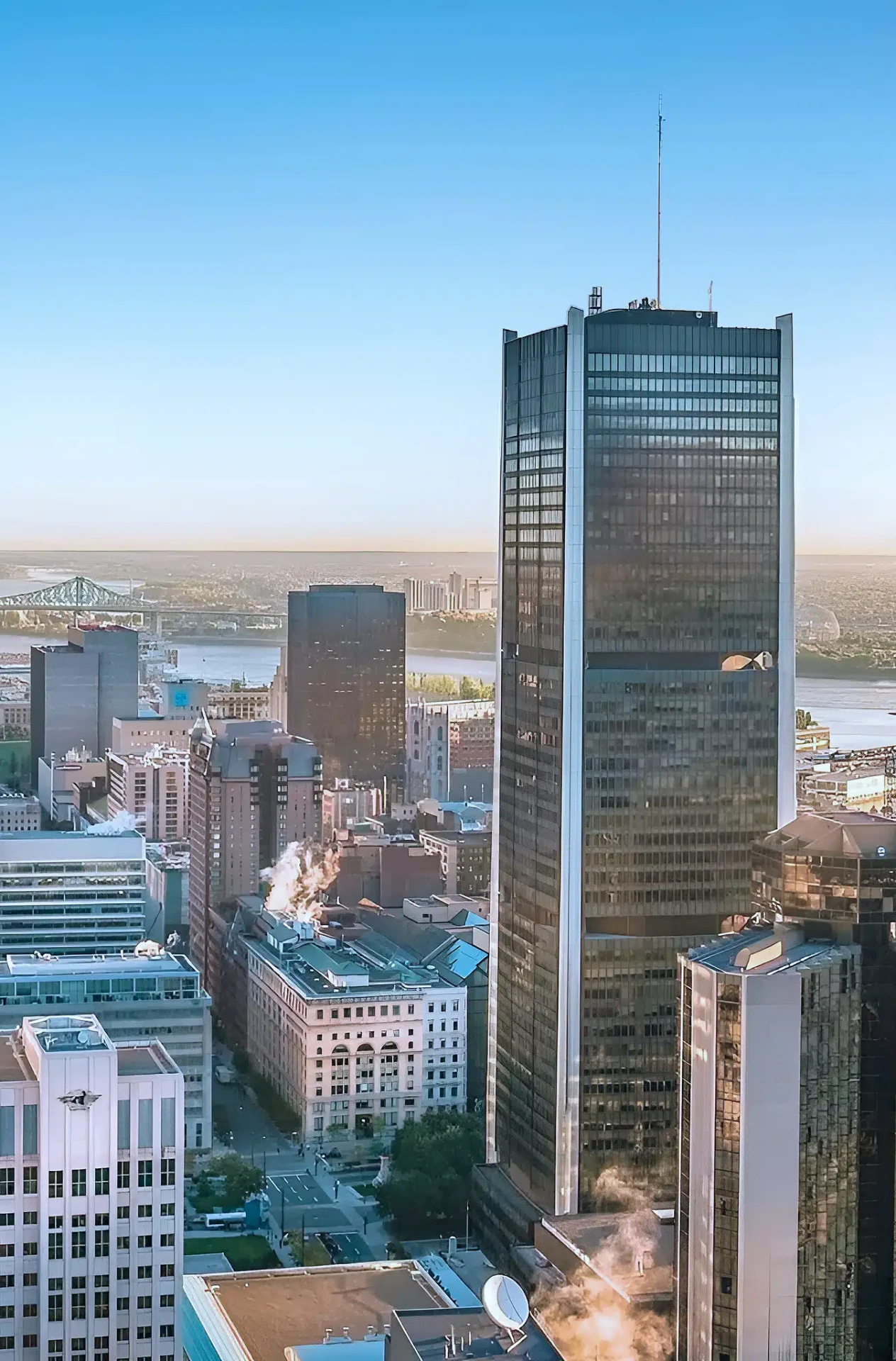 Downtown Montréal skyline at golden hour: a tower at Place Victoria, with the Jacques Cartier Bridge and the Saint Lawrence River in the distance